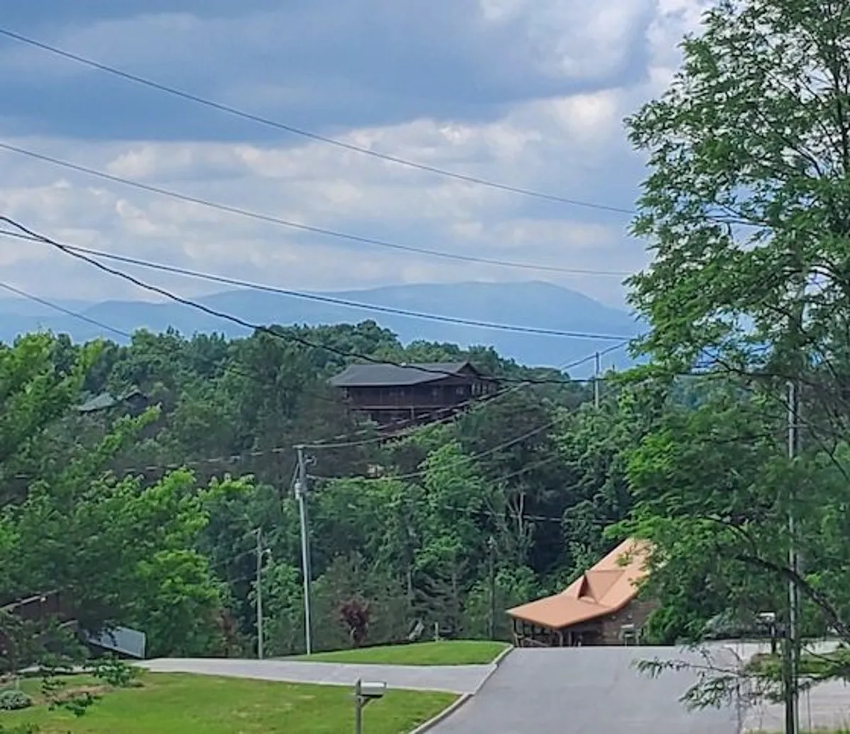 A wide, panoramic view of the Smoky Mountains with lush greenery and a clear blue sky. A winding road leads up to a charming house with a distinctive roof, nestled amidst nature. The perspective hints at exploration and proximity to local attractions and scenic drives.