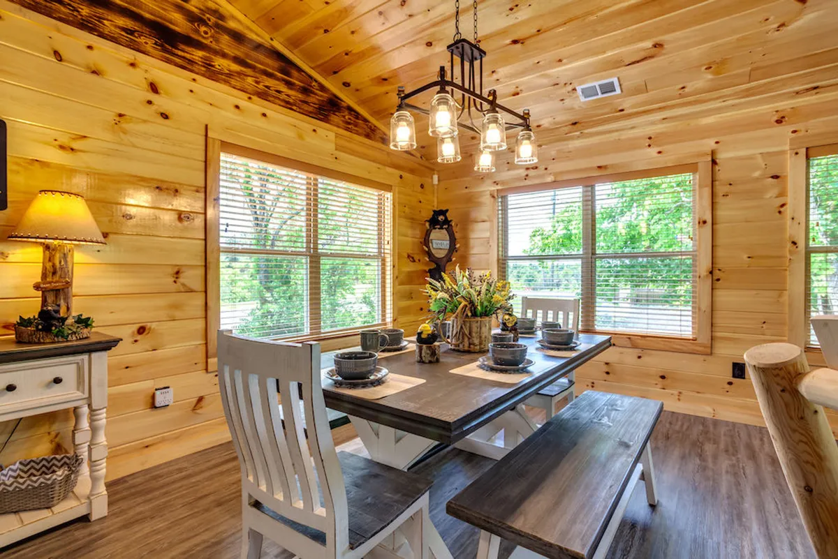 An elegant dining area featuring a wooden table under a chandelier, surrounded by windows that let in natural light. A side lamp and decorative vase add a touch of charm, suggesting a perfect setting for family meals or celebrations.
