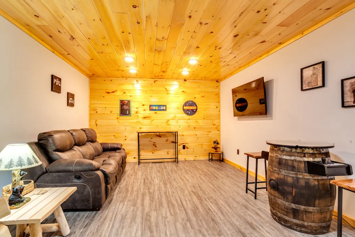 The relaxed den setup includes a reclining sofa, wooden barrel table positioned for board or card games, and a wall-mounted TV. Framed pictures add a personal touch, emphasizing a space designed for fun and togetherness.