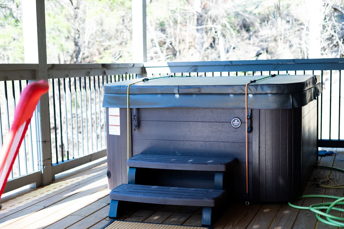 A view of a hot tub with steps leading up to it on an outdoor deck, set against a backdrop of natural greenery. The scene is tranquil, suggesting a secluded space for soaking while enjoying the outdoors.
