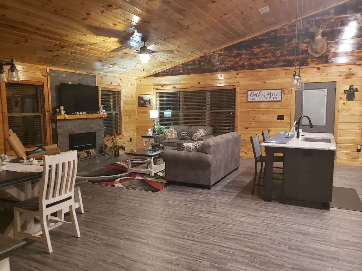 A spacious open-concept living and dining area. The image showcases a comfortable sofa, a dining table set for multiple guests, a cozy fireplace, a wall-mounted TV, and a kitchen island in the background. Warm lighting and wood accents create a welcoming atmosphere ideal for gatherings.