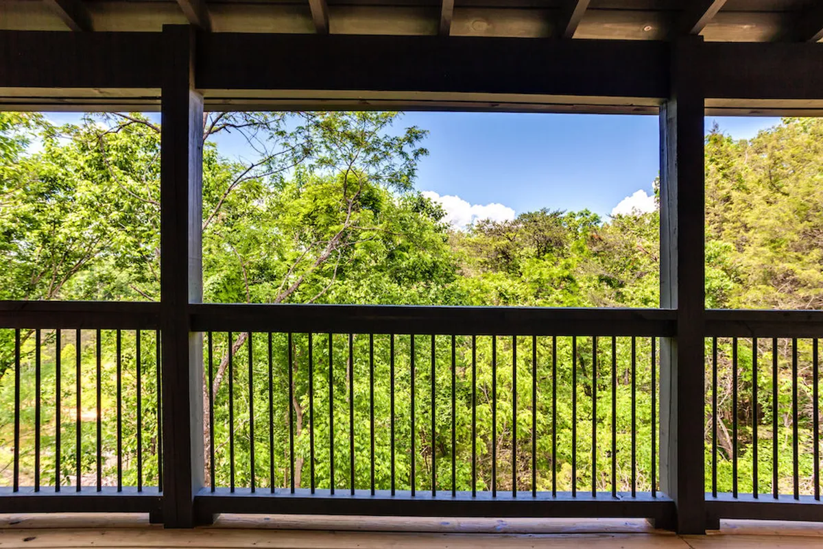 A serene view from a balcony or deck, overlooking green foliage and a blue sky, framed by a rustic wooden railing. The setting invites quiet reflection, with the feeling of being immersed in nature. This is an ideal spot for sipping a morning coffee, reading, or simply enjoying the mountain views.
