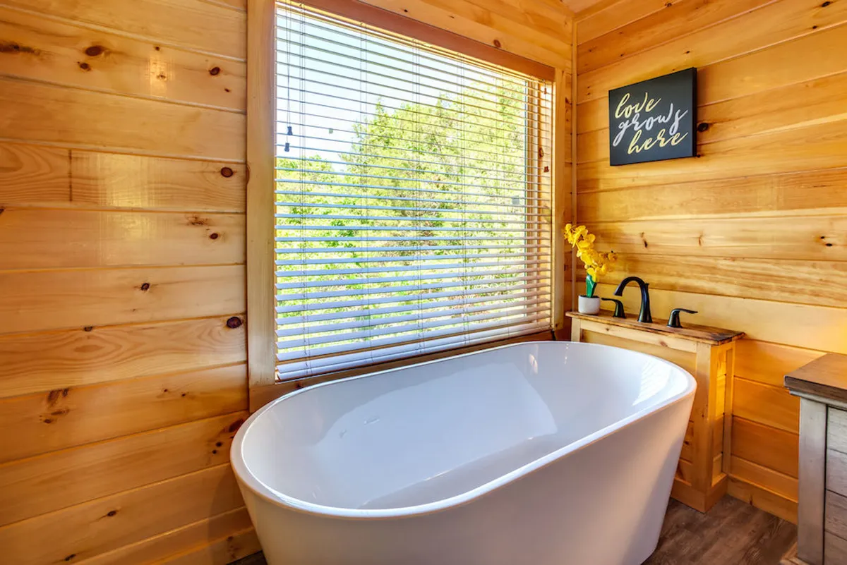 A serene, spa-like bathroom with a freestanding bathtub, window with blinds for privacy, wooden walls, and a decorative sign, evoking a relaxing, rejuvenating atmosphere.