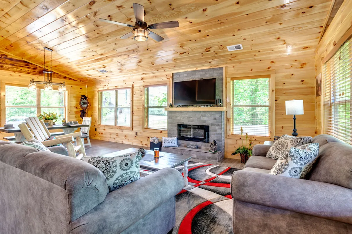 The image showcases a warm and spacious open-concept living area featuring a soaring wooden ceiling, plush sofas gathered around a stone fireplace, a family dining area, and expansive windows that frame serene mountain views. Natural light fills the space, creating an inviting atmosphere perfect for cozy evenings and lively gatherings.